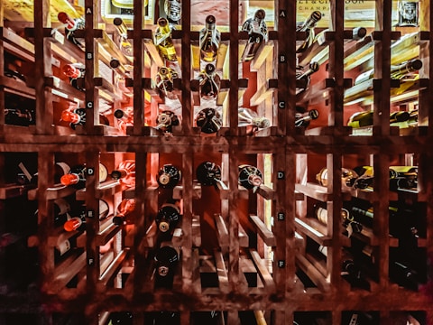 A well-organized wine rack filled with numerous bottles of wine, arranged horizontally and neatly labeled with letters. Various shades of red, brown, and amber are visible on the necks and bodies of the bottles. Soft lighting casts shadows and highlights the textures of the wooden rack.