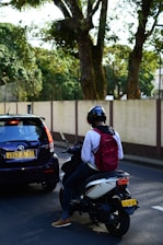 A person wearing a helmet and a red backpack is sitting on a scooter next to a black car on a road. The car and scooter are both stopped, with the car's license plate visible. The background features a fence and large trees with dense foliage.