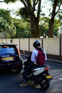 A person wearing a helmet and a red backpack is sitting on a scooter next to a black car on a road. The car and scooter are both stopped, with the car's license plate visible. The background features a fence and large trees with dense foliage.