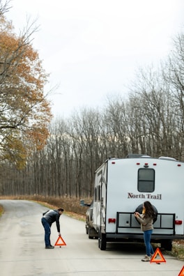 A man and a woman are on a road beside a parked RV. The man is setting up an orange warning triangle on the ground, while the woman stands nearby holding a phone. The scene is surrounded by a forest with trees mostly bare of leaves, indicating a fall or winter setting.