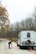 A man and a woman are on a road beside a parked RV. The man is setting up an orange warning triangle on the ground, while the woman stands nearby holding a phone. The scene is surrounded by a forest with trees mostly bare of leaves, indicating a fall or winter setting.