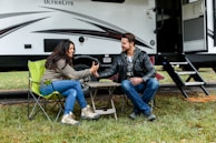 A man and a woman are sitting on camping chairs outside a travel trailer, smiling and holding hands across a small folding table. The atmosphere appears relaxed and friendly in a natural setting with grass and fallen leaves.