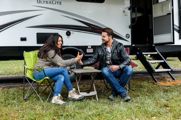 A man and a woman are sitting on camping chairs outside a travel trailer, smiling and holding hands across a small folding table. The atmosphere appears relaxed and friendly in a natural setting with grass and fallen leaves.