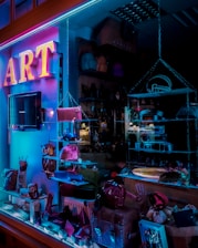 A store display window features various accessories, including purses and handbags, under vibrant blue and pink neon lighting. The interior of the store is dimly lit, highlighting the colorful and artistic presentation of the products.