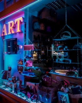 A store display window features various accessories, including purses and handbags, under vibrant blue and pink neon lighting. The interior of the store is dimly lit, highlighting the colorful and artistic presentation of the products.