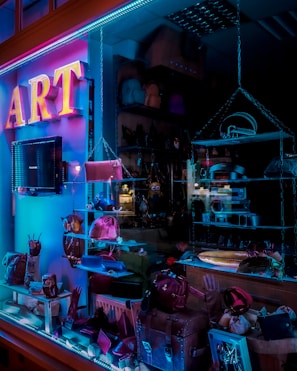 A store display window features various accessories, including purses and handbags, under vibrant blue and pink neon lighting. The interior of the store is dimly lit, highlighting the colorful and artistic presentation of the products.