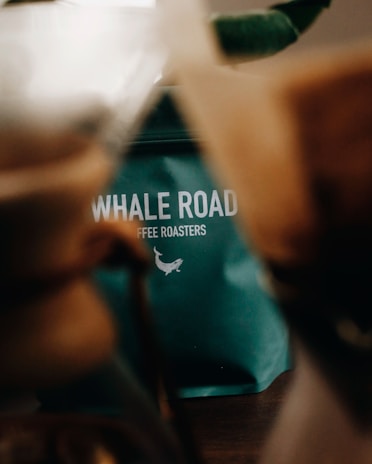 A teal-green coffee bag with the text 'WHALE ROAD COFFEE ROASTERS' partially visible, blurred out objects in the foreground suggest a coffee-making setup.