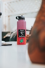 A red water bottle with a black cap is placed on a white table. The bottle features a black sticker with the words 'Social Distancing' and a graphic image of a skeleton. A hand gesture with a green glove is illustrated below the text. In the background, parts of another water bottle and a blurred figure are visible.