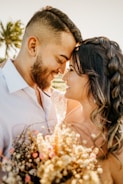 A couple is sharing an intimate moment, with their foreheads touching and eyes closed. They are smiling warmly at each other, surrounded by warm sunlight and soft focus, which adds to the romantic atmosphere. The woman is holding a bouquet of dried flowers with subtle pink and cream hues. In the background, a palm tree and gently blurred scenery add to the calm and serene setting.