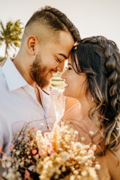 A couple is sharing an intimate moment, with their foreheads touching and eyes closed. They are smiling warmly at each other, surrounded by warm sunlight and soft focus, which adds to the romantic atmosphere. The woman is holding a bouquet of dried flowers with subtle pink and cream hues. In the background, a palm tree and gently blurred scenery add to the calm and serene setting.
