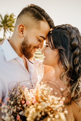A couple is sharing an intimate moment, with their foreheads touching and eyes closed. They are smiling warmly at each other, surrounded by warm sunlight and soft focus, which adds to the romantic atmosphere. The woman is holding a bouquet of dried flowers with subtle pink and cream hues. In the background, a palm tree and gently blurred scenery add to the calm and serene setting.