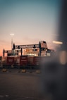 A transport terminal features several red freight containers lined up under an industrial structure with blue steel frameworks. A ferry or large ship is partially visible behind them, docked and ready for loading. The sky is at dusk, transitioning from a soft pink near the horizon to a deeper blue-gray higher up. Bright artificial lighting in the scene creates a contrast with the natural twilight.