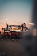 A transport terminal features several red freight containers lined up under an industrial structure with blue steel frameworks. A ferry or large ship is partially visible behind them, docked and ready for loading. The sky is at dusk, transitioning from a soft pink near the horizon to a deeper blue-gray higher up. Bright artificial lighting in the scene creates a contrast with the natural twilight.