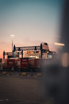 A transport terminal features several red freight containers lined up under an industrial structure with blue steel frameworks. A ferry or large ship is partially visible behind them, docked and ready for loading. The sky is at dusk, transitioning from a soft pink near the horizon to a deeper blue-gray higher up. Bright artificial lighting in the scene creates a contrast with the natural twilight.