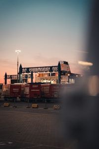 A transport terminal features several red freight containers lined up under an industrial structure with blue steel frameworks. A ferry or large ship is partially visible behind them, docked and ready for loading. The sky is at dusk, transitioning from a soft pink near the horizon to a deeper blue-gray higher up. Bright artificial lighting in the scene creates a contrast with the natural twilight.