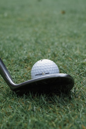 A close-up of a golf ball positioned on grass near the head of a golf club, with the text 'VERO X1' visible on the ball.