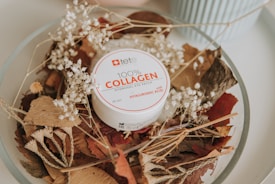 A round container labeled '100% COLLAGEN Hydrogel Eye Patch' is placed on a bed of dried leaves and white flowers. The container is white with red and black text, and the surrounding leaves vary in shades of brown and beige, creating a natural and organic aesthetic. A light blue object is partially visible in the background, adding a subtle contrast.