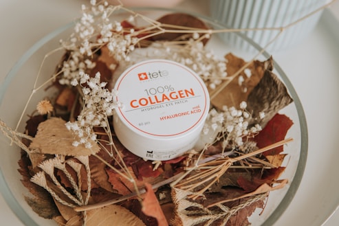 A round container labeled '100% COLLAGEN Hydrogel Eye Patch' is placed on a bed of dried leaves and white flowers. The container is white with red and black text, and the surrounding leaves vary in shades of brown and beige, creating a natural and organic aesthetic. A light blue object is partially visible in the background, adding a subtle contrast.
