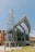 A modern architectural structure featuring a cylindrical tower with a crisscross metal design and a large, curved glass facade. The building is surrounded by greenery and a clear blue sky, giving it a contemporary and open feel.