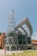 A modern architectural structure featuring a cylindrical tower with a crisscross metal design and a large, curved glass facade. The building is surrounded by greenery and a clear blue sky, giving it a contemporary and open feel.
