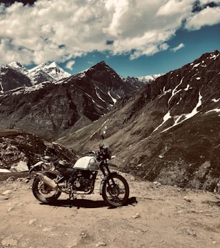 A motorcycle is parked on a rocky, mountainous terrain under a partly cloudy sky. Snow-capped peaks and rugged slopes dominate the background, indicating a high-altitude environment. The motorcycle appears to be an adventure or off-road type, suitable for navigating such rough terrains.