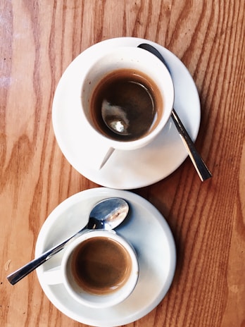 Two white cups of espresso sit on saucers on a wooden table. Each cup has a metal spoon resting on the saucer, and the espresso is dark and rich-looking.