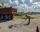 A person wearing a reflective orange vest is working on patching a road surface. They are using a tool to spread asphalt on a small section of the road. A large dump truck is parked nearby, likely used for transporting materials. The surrounding landscape is lush with greenery, and there are fields with water, possibly rice paddies, under a partly cloudy sky.