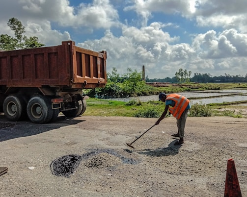 A person wearing a reflective orange vest is working on patching a road surface. They are using a tool to spread asphalt on a small section of the road. A large dump truck is parked nearby, likely used for transporting materials. The surrounding landscape is lush with greenery, and there are fields with water, possibly rice paddies, under a partly cloudy sky.