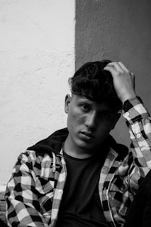 A person with a thoughtful expression leans against a textured wall. They have short, wavy hair and are wearing a checkered flannel over a dark shirt. Their hand is placed on top of their head, adding to the introspective mood.