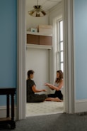 Two people are seated on the floor in a small room with soft blue walls, carrying out a focused interaction. Natural light enters through a tall window. The floor is light-colored with small dark patterns. Above them, some boxes and books are stored on a shelf. A decorative ceiling light fixture is visible.