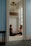Two people are seated on the floor in a small room with soft blue walls, carrying out a focused interaction. Natural light enters through a tall window. The floor is light-colored with small dark patterns. Above them, some boxes and books are stored on a shelf. A decorative ceiling light fixture is visible.