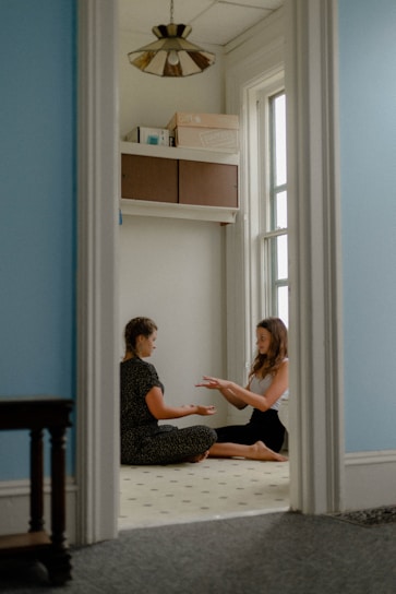 Two people are seated on the floor in a small room with soft blue walls, carrying out a focused interaction. Natural light enters through a tall window. The floor is light-colored with small dark patterns. Above them, some boxes and books are stored on a shelf. A decorative ceiling light fixture is visible.