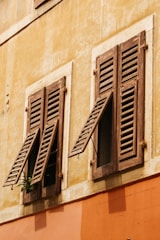 A rustic building facade features two old wooden shutters, slightly open, mounted on a weathered yellow and orange wall. A small plant peeks through one of the shutters, adding a touch of greenery to the aged surface.