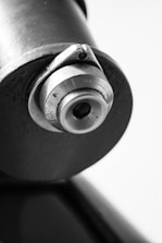 A close-up of a metallic cylinder with a textured, industrial appearance. The focus is on a circular component featuring ridges and a valve-like structure.