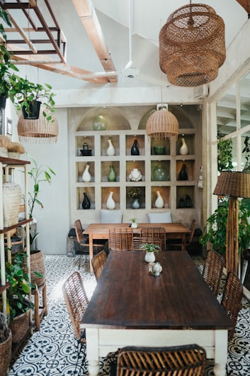 A cozy dining area with wooden furniture and wicker chairs is adorned with hanging and potted plants. The floor features intricate patterned tiles, and shelves on the wall display various decorative vases, mostly in neutral and earthy tones. Natural light streams in through an unseen source.