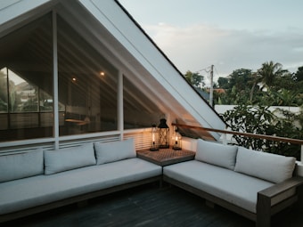 An outdoor patio area with a modern design, featuring two gray sectionals creating an L-shape around a small wooden table. Lanterns with candles are placed on the table, emitting a warm, cozy light. The area is enclosed by a wooden railing with lush greenery and palm trees visible in the background. The architecture includes a large glass window with a triangular frame, showcasing a glimpse of the interior structure.