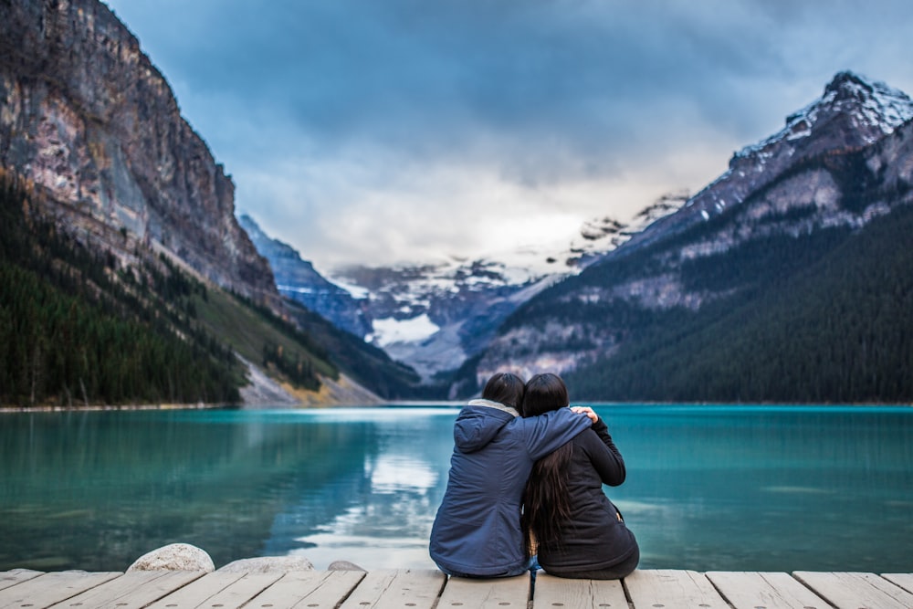 man in black jacket and blue denim jeans sitting on brown wooden dock looking at the near near near near