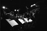 A monochrome photograph of a group of musicians playing brass instruments, possibly trombones, while reading sheet music on stands in a dimly lit venue. The background is dark and the overhead stage lights create a dramatic atmosphere, illuminating some parts of the image.