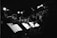 A monochrome photograph of a group of musicians playing brass instruments, possibly trombones, while reading sheet music on stands in a dimly lit venue. The background is dark and the overhead stage lights create a dramatic atmosphere, illuminating some parts of the image.