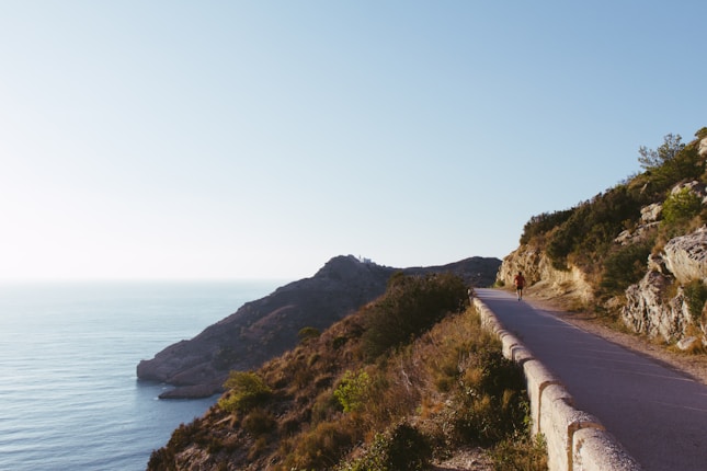 A scenic coastal road runs alongside a rocky hillside with dense greenery, leading towards the ocean. A solitary cyclist wearing red rides along the road, under a clear blue sky. The landscape captures a tranquil meeting of land and sea, with distant hills and the vast expanse of the water.