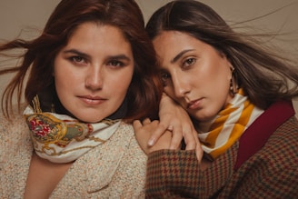 Two people pose closely together, both wearing stylish scarves and coats. One has auburn hair and wears a scarf with a detailed pattern, while the other has long dark hair and wears a striped scarf.