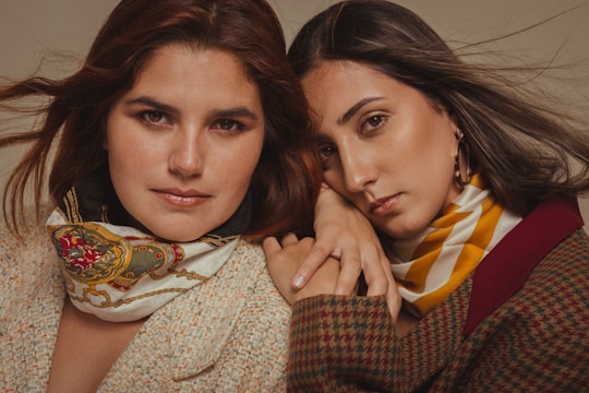 Two people pose closely together, both wearing stylish scarves and coats. One has auburn hair and wears a scarf with a detailed pattern, while the other has long dark hair and wears a striped scarf.