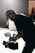 A person wearing a cap is operating a professional video camera. The scene is dimly lit, with a soft light source creating a dramatic silhouette effect. The background is blurred, suggesting a studio or indoor environment.