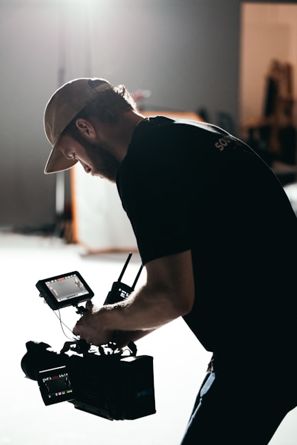 A person wearing a cap is operating a professional video camera. The scene is dimly lit, with a soft light source creating a dramatic silhouette effect. The background is blurred, suggesting a studio or indoor environment.