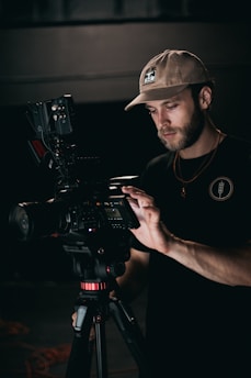 A man stands focused on operating a professional video camera mounted on a tripod. He wears a beige cap and a dark shirt, and carefully adjusts settings on the camera. The lighting is low, emphasizing the concentration and precision of his work.