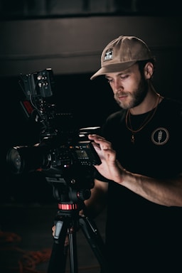 A man stands focused on operating a professional video camera mounted on a tripod. He wears a beige cap and a dark shirt, and carefully adjusts settings on the camera. The lighting is low, emphasizing the concentration and precision of his work.
