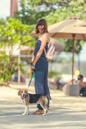 A woman in a blue polka dot jumpsuit stands outdoors on a sunny day, holding a leash connected to a beagle dog. She is smiling and carrying a small backpack. The background features green trees and large beige umbrellas providing shade.