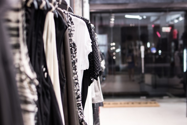 A row of assorted clothing hanging on a rack, with a focus on various textures and patterns. Neutral tones dominate, with blacks, whites, and grays prominently displayed. The setting appears to be inside a modern retail store with glass windows reflecting lights and other store details in the background.