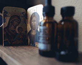 A religious icon depicting a figure with a halo and crown, likely representing a holy person, is placed on a wooden surface. In the foreground, several amber-colored glass bottles are slightly out of focus.
