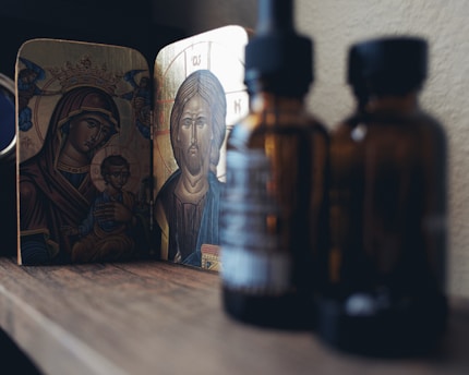 A religious icon depicting a figure with a halo and crown, likely representing a holy person, is placed on a wooden surface. In the foreground, several amber-colored glass bottles are slightly out of focus.